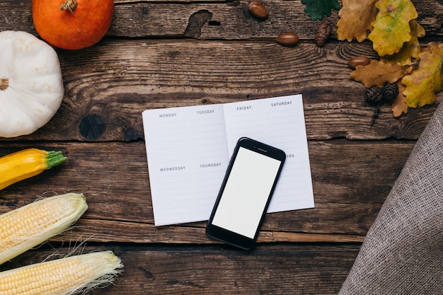 Verduras de otoño: teléfono móvil con pantalla blanca vacía, calabazas y maíz con hojas amarillas en madera