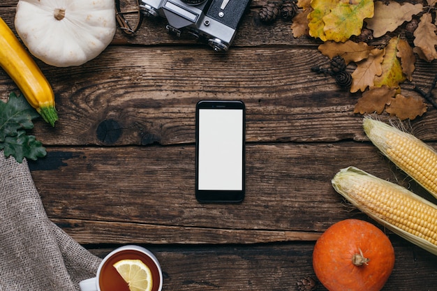 Verduras de otoño: teléfono móvil con pantalla blanca vacía, calabazas y maíz con hojas amarillas en madera