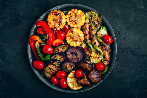 Verduras a la parrilla en un tazón sobre fondo de piedra oscura vista superior Zucchini pimientos maíz dulce tomates cebolla roja y berenjena