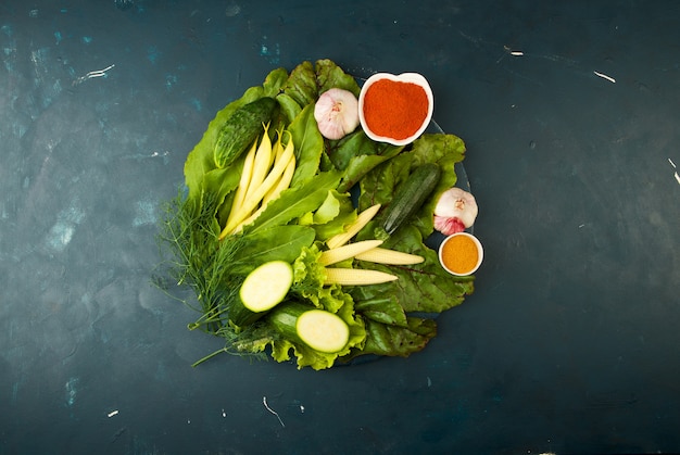 VERDURAS EN EL TABLERO DE MADERA EN UNA PIEDRA. JOVENES HIERBAS CEBOLLAS AJO HABAS VERDES MAÍZ AMARILLO ZUCCHINI ESPECIAS BRILLANTES MIENTEN EN UN TABLERO DE MADERA OSCURO EN UN TEXTURADO.