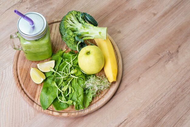 Verduras verdes frescas y batido verde en frasco. Concepto de desintoxicación, dieta o comida sana. Mason jar de bebida dietética con brócoli, espinacas, microgreens, limón y plátano sobre fondo de madera. Vista superior.