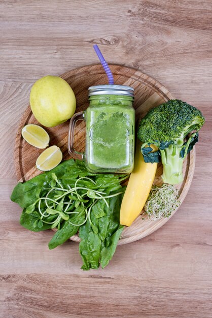 Verduras verdes frescas y batido verde en frasco. Concepto de desintoxicación, dieta o comida sana. Mason jarra de bebida dietética con brócoli, espinacas, microgreens, limón y plátano en la mesa de madera. Vista superior.