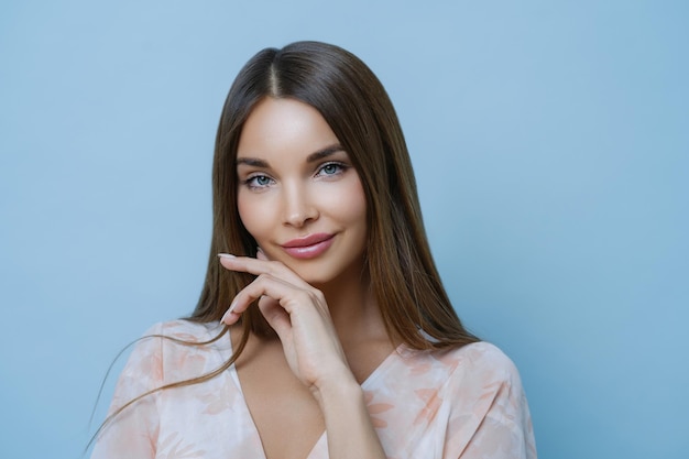 Verführerische, angenehm aussehende Frau mit langen, dunklen, glatten Haaren berührt die Kinnpartie und freut sich auf ein besonderes Ereignis, das an einem professionellen Fotoshooting teilnimmt und gesunde, saubere Haut auf blauem Hintergrund posiert