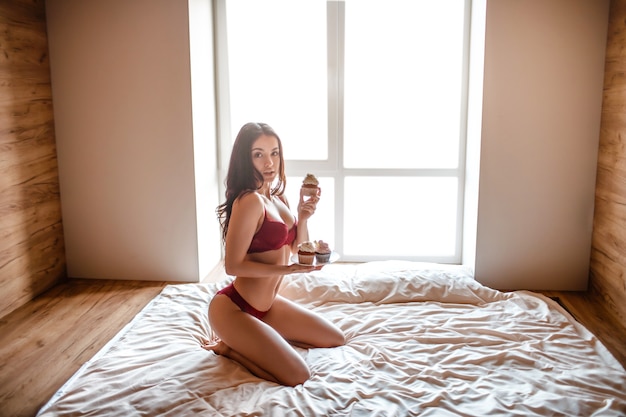 Verführerische junge nackte dunkelhaarige Frau morgens auf dem Bett. Sexy Model mit Pfannkuchen auf Teller in den Händen und Blick in die Kamera. Sinnliche Pose am Fenster