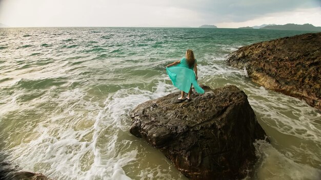 Verführerische langhaarige Frau geht auf Felsen des Meeresriffsteins stürmischer bewölkter Ozean Frau in blauem Swimsui