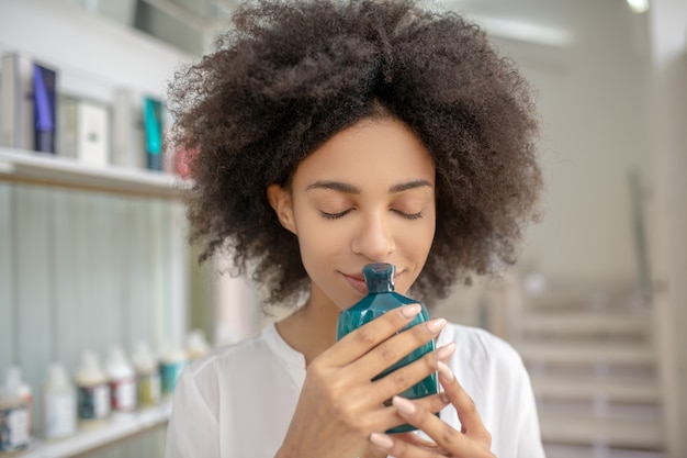 Vergnügen, Aroma. Gesicht des hübschen Mädchens mit dunklem lockigem Haar mit geschlossenen Augen, die Aroma von Flasche in Händen einatmen