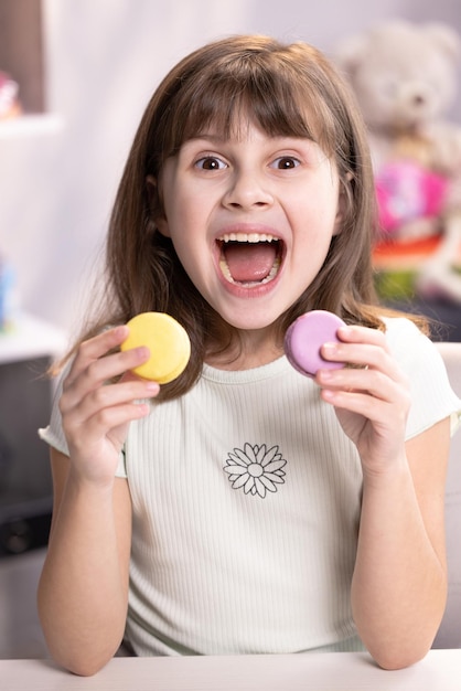 Vergnügtes Schulmädchen will gleichzeitig zwei Macarons essen, öffnet weit ihren Mund Nachtisch