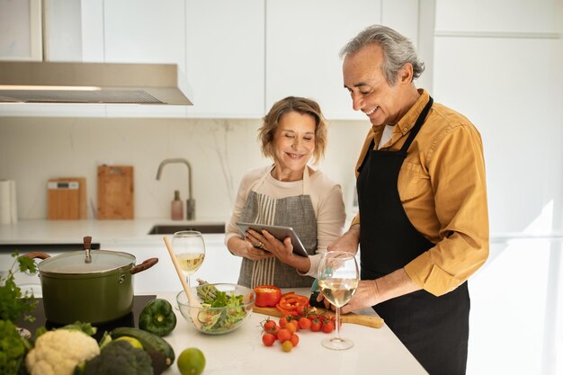 Verheiratetes älteres Paar, das gemeinsam in der Küche Essen kocht und ältere Ehepartner mit digitalen Tablets verwendet