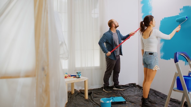 Verheiratetes Paar, das während der Hausrenovierung mit blauer Farbe die Wohnungswand mit Walze streicht. Dekorieren, Farbe, Reparatur, Dekoration.