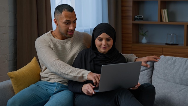 Verheiratetes Paar sitzt auf dem Sofa und benutzt ein Laptop-Gerät, das eine neue Wohnung auf der multirassischen Internet-Website auswählt
