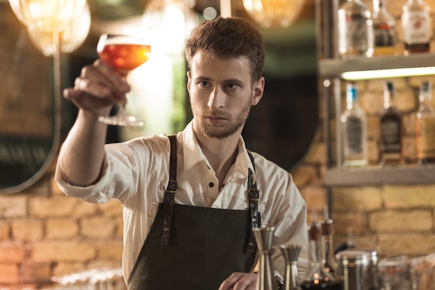 Verificação completa. Bonito barman profissional erguendo um copo com um coquetel e olhando atentamente para ele enquanto verifica a cor da bebida