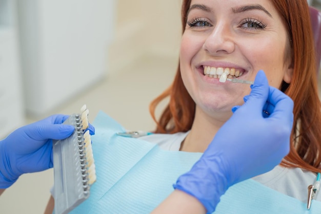 Verifique e selecione a cor dos dentes na cadeira do dentista Dentista faz o processo de tratamento de uma bela jovem ruiva Seleção de um implante dentário Closeup