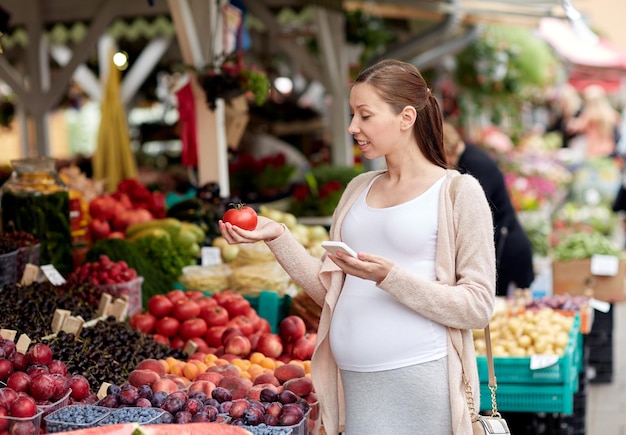 Verkauf, Einkaufen, Essen, Schwangerschaft und Personenkonzept - glückliche schwangere Frau mit Smartphone, die Gemüse auf dem Straßenmarkt auswählt