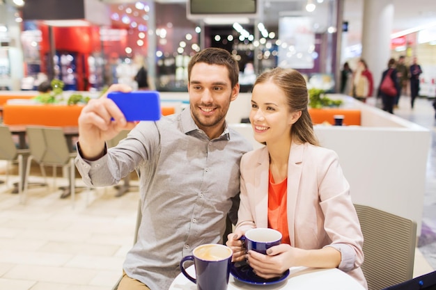 verkauf, einkaufen, konsum, technologie und menschenkonzept - glückliches junges paar mit smartphone, das selfie im café im einkaufszentrum macht