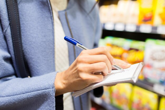 verkauf, einkaufen, konsum und personenkonzept - nahaufnahme einer jungen frau mit stift, die sich notizen zum notizbuch auf dem markt macht