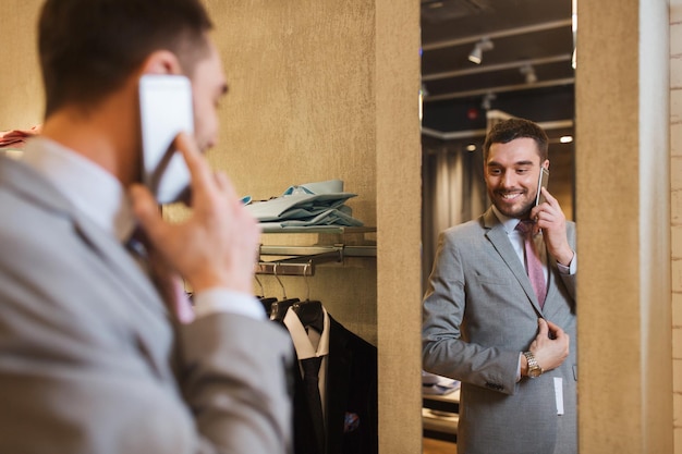 Verkauf, Einkaufen, Mode, Stil und People-Konzept - glücklicher junger Mann oder Geschäftsmann, der Anzug und Krawatte anprobiert und Smartphone im Spiegel des Bekleidungsgeschäfts anruft