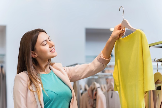 verkauf, einkaufen, mode, stil und personenkonzept - glückliche junge frau, die kleidung in einem einkaufszentrum oder einem bekleidungsgeschäft auswählt