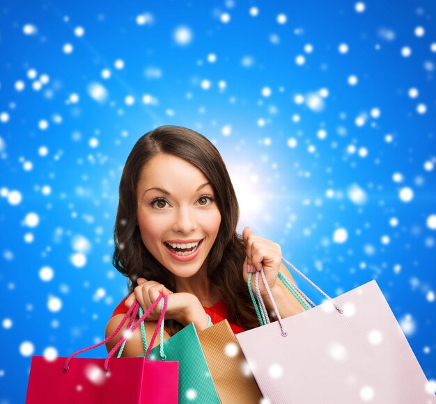 verkauf, geschenke, weihnachten, feiertage und personenkonzept - lächelnde frau mit bunten einkaufstüten über blauem schneebedecktem hintergrund
