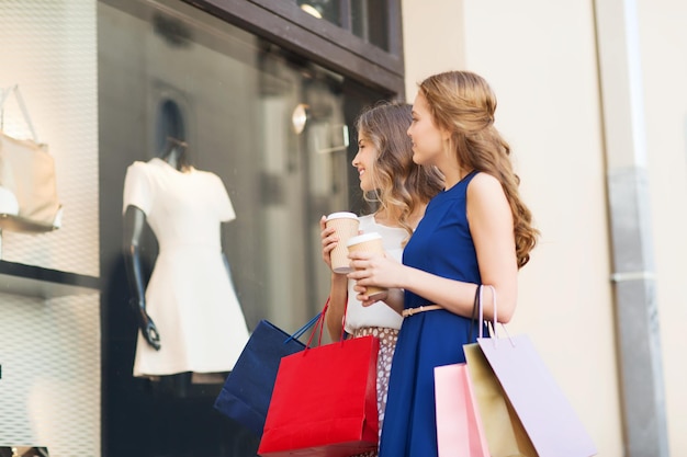 Verkauf, Konsum und Menschenkonzept - glückliche junge Frauen mit Einkaufstüten und Kaffeepappbechern, die im Freien auf das Schaufenster schauen