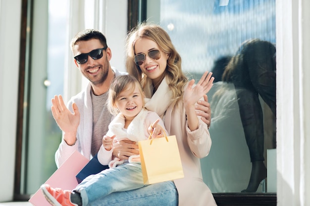verkauf, konsum und personenkonzept - glückliche familie mit kleinem kind und einkaufstüten winken am schaufenster in der stadt