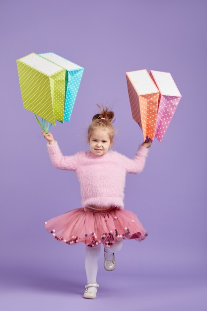 Verkauf von kinderprodukten. porträt des lustigen kleinen kindermädchens auf lila raum, der einkaufstaschen, paket hält. blick in die kamera. banner. platz für text.