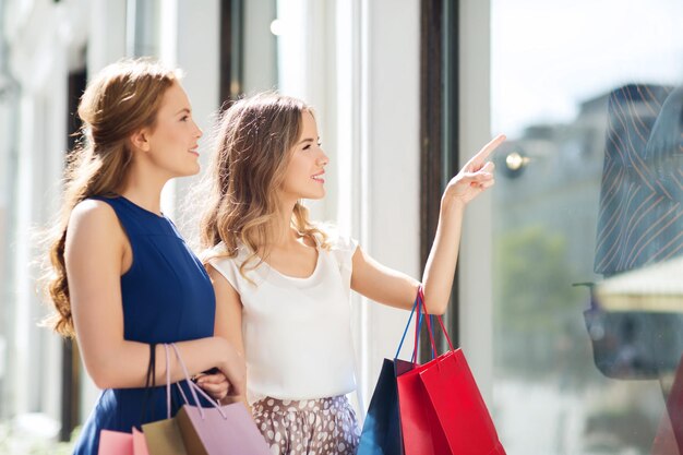 Foto verkaufs-, konsum- und menschenkonzept - glückliche junge frauen mit einkaufstüten, die mit dem finger auf das schaufenster in der stadt zeigen