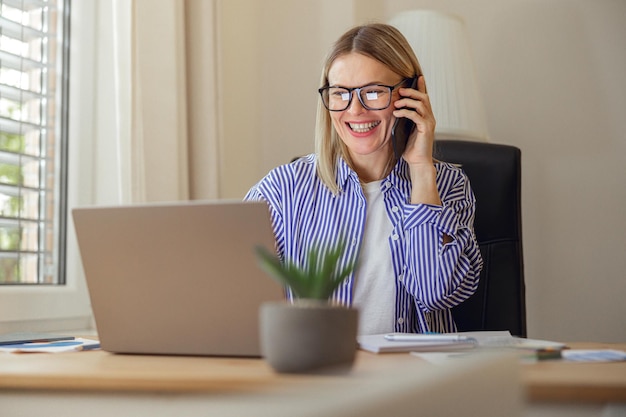 Verkaufsleiterin mit Brille spricht am Telefon mit einem Kunden, der von zu Hause aus am Laptop arbeitet
