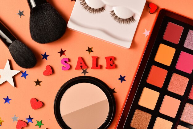 Verkaufstext auf einem orange Hintergrund. Professionelle trendige Make-up-Produkte mit kosmetischen Schönheitsprodukten, Lidschatten, Wimpern, Pinseln und Werkzeugen.