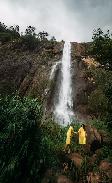 Verliebtes Paar am Wasserfall. Jungen und Mädchen an den Wasserfällen. Mann und Frau in gelben Regenmänteln. Kerl und Mädchen reisen. Das Paar reist durch Asien. Wasserfälle in Sri Lanka. Flitterwochen