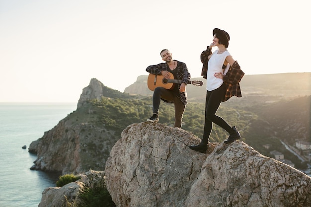 Verliebtes Paar auf einem Berggipfel mit Blick aufs Meer Mann spielt Gitarre, eine Frau tanzt und singt
