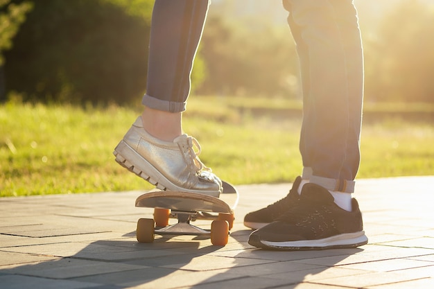 Verliebtes Paar in einem Sommerpark. Beine Füße eines Mannes und einer Frau in modischen Jeans und stylischen Turnschuhen auf einem Skateboard-Longboard-Fußweg-Asphalt. Jugendaktivität und das erste Liebeskonzept.