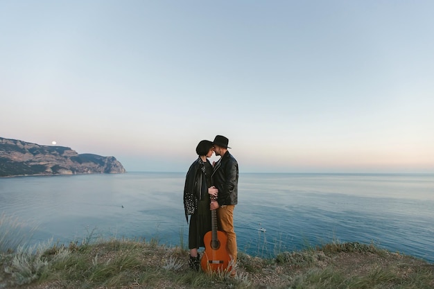 Verliebtes Paar mit einer Gitarre auf dem Gipfel des Berges mit Blick auf das Meer Mann und Frau umarmen sich bei Sonnenuntergang
