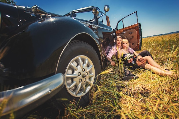 Verliebtes Paar sitzt in der Nähe eines alten schwarzen Autos auf einem Weizenfeld