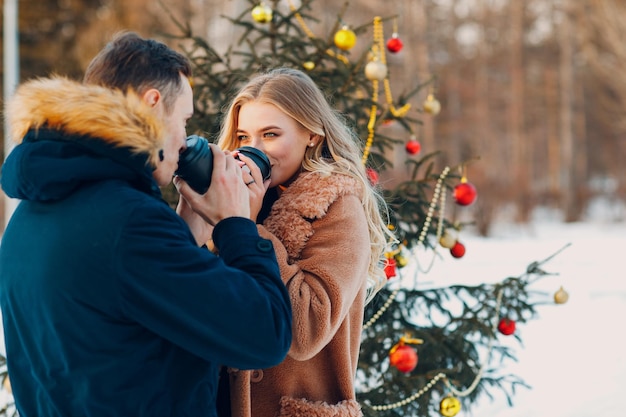 Verliebtes Paar trinkt Kaffee neben geschmücktem Weihnachtsbaum im Winterwald.