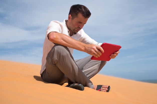 Verloren in der Wüste Kaukasischer Mann, der auf Sand mit Tablet sitzt. Er ist verloren und sucht auf der Karte, wohin er gehen soll