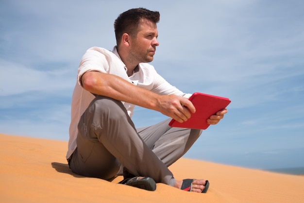 Verloren in der Wüste Kaukasischer Mann, der auf Sand mit Tablet sitzt. Er ist verloren und sucht auf der Karte, wohin er gehen soll