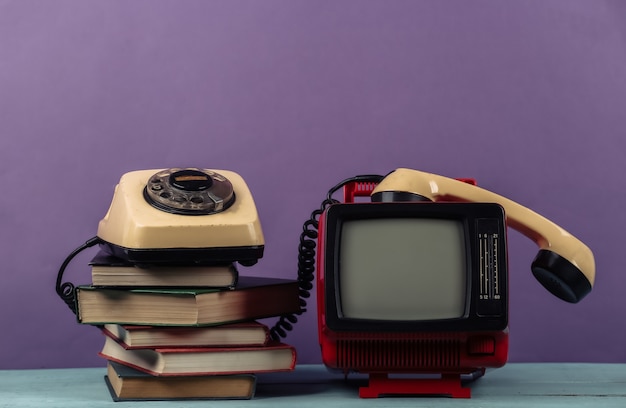 Vermelho retro velho mini tv portátil com telefone giratório, pilha de livros sobre fundo roxo.