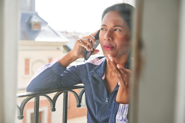 Vernetzt und produktiv Junge indische Frau multitaskt auf ihrem Telefon, während sie auf der Fensterbank sitzt