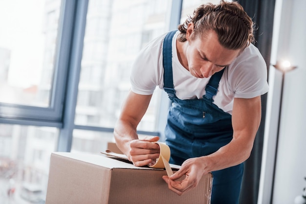 Verpacken der Box Junger männlicher Umzugshelfer in blauer Uniform arbeitet drinnen im Raum