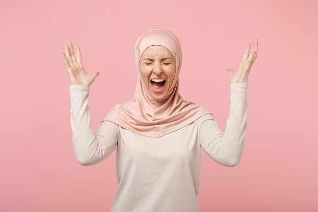 Verrückte junge arabische muslimische Frau in Hijab-leichter Kleidung posiert isoliert auf rosa Hintergrund. Menschen religiösen Islam Lifestyle-Konzept. Mock-up-Kopienbereich. Schreien, Augen geschlossen halten, Hände ausbreiten.