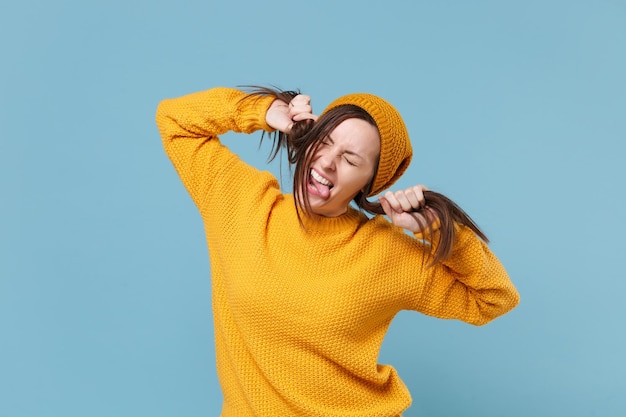 Verrückte junge brünette Frau Mädchen in gelbem Pullover Hut posiert isoliert auf blauem Hintergrund Studioportrait. Menschen aufrichtige Emotionen Lifestyle-Konzept. Mock-up-Kopienbereich. Haare wie Pferdeschwänze halten.