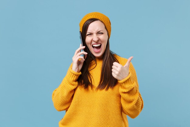 Verrückte junge Frau in gelbem Pullover und Hut posiert isoliert auf blauem Hintergrund Studioportrait. Menschen aufrichtige Emotionen Lifestyle-Konzept. Mock-up-Kopienbereich. Telefonieren mit dem Daumen nach oben.