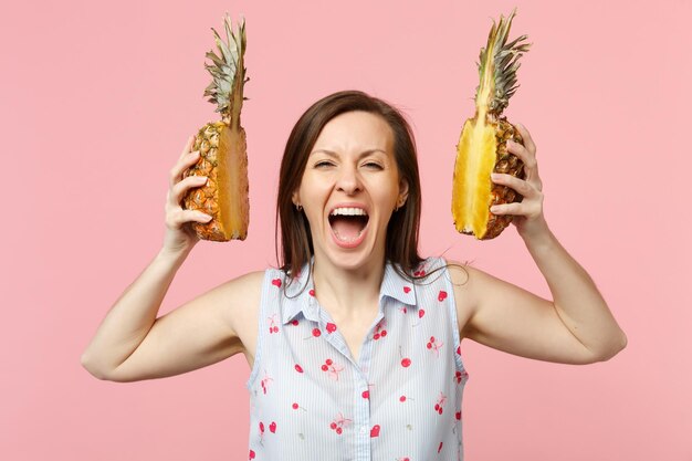 Verrückte schreiende junge Frau in Sommerkleidung hält Hälften frischer reifer Ananasfrüchte isoliert auf rosa Pastellhintergrund im Studio. Menschen lebendiger Lebensstil entspannen Urlaubskonzept. Mock-up-Kopienbereich.