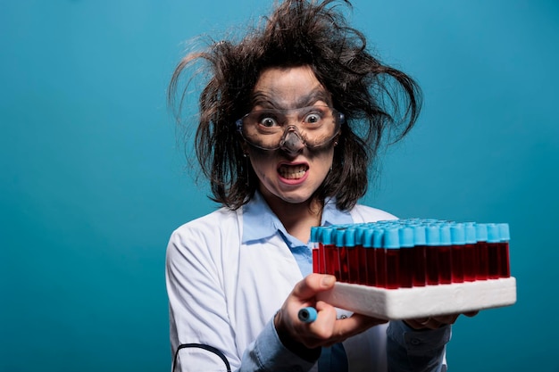 Verrückter und wütender Chemiker mit Laborglasschale mit Reagenzgläsern, die mit roter flüssiger Substanz gefüllt sind. Wahnsinniger wütender Wissenschaftler, der Laborglasröhrchen mit Proben auf blauem Hintergrund gefüllt hat.