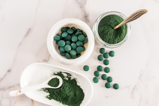 Verschiedene Behälter gefüllt mit grünem Algenpulver Spirulina und Tabletten auf weißem Marmorhintergrund hoher Gehalt an Multivitaminen Detox