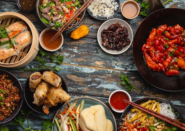 Verschiedene chinesische Gerichte auf dem Tisch: süßes und saures Hühnchen in der Wok-Pfanne, Dim Sum, Frühlingsrollen, Nudeln, Salat, Reis, gedämpfte Brötchen. Abendessen oder Buffet im asiatischen Stil mit Draufsicht und Platz für Text