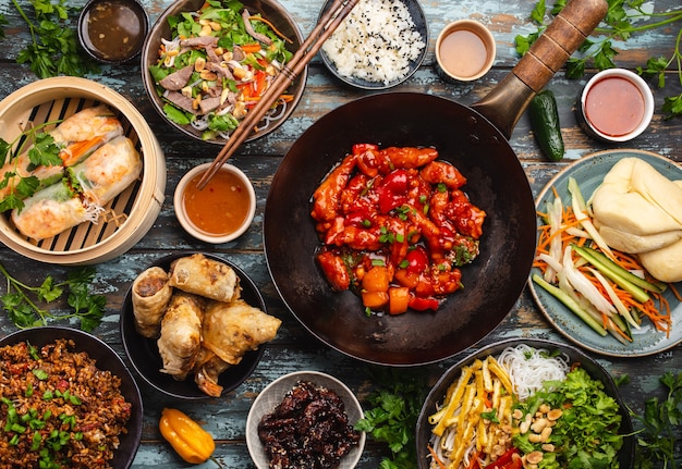 Verschiedene chinesische Gerichte auf dem Tisch: süßes und saures Hühnchen in der Wok-Pfanne, Dim Sum im Bambusdampfer, Frühlingsrollen, Nudeln, Salat, Reis, gedämpfte Brötchen, Dips. Abendessen oder Buffet im asiatischen Stil, Draufsicht