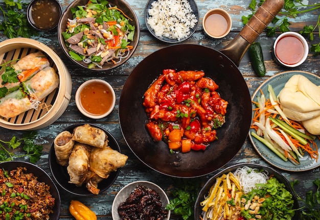 Verschiedene chinesische Gerichte auf dem Tisch: süßes und saures Hühnchen in der Wok-Pfanne, Dim Sum im Bambusdampfer, Frühlingsrollen, Nudeln, Salat, Reis, gedämpfte Brötchen, Dips. Abendessen oder Buffet im asiatischen Stil, Draufsicht