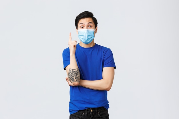 Verschiedene Emotionen, soziale Distanzierung, Selbstquarantäne bei Coronavirus und Lifestyle-Konzept. Aufgeregter asiatischer Typ in blauem T-Shirt und medizinischer Maske, heben Sie den Zeigefinger an, haben Sie eine Lösung, eine Idee?