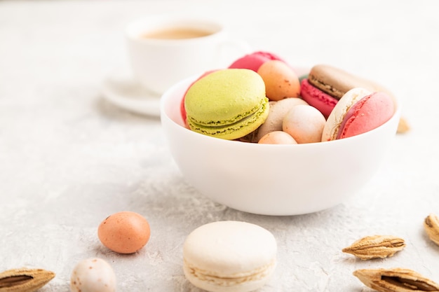 Verschiedene Farben Makronen und Schokoladeneier in Keramikschale Tasse Kaffee auf grauem Betonhintergrund Seitenansicht Nahaufnahme